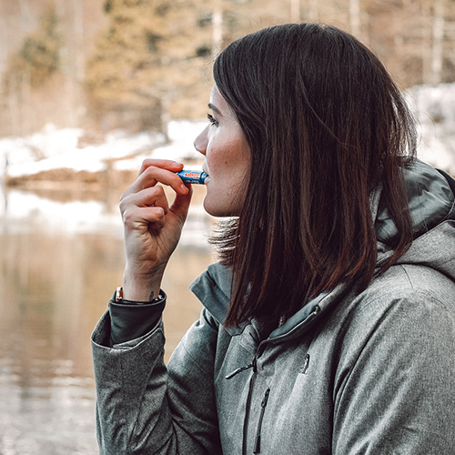 Eine Frau benutzt ein Blistex-Lippenprodukt und steht an einem See in verschneiter Landschaft.