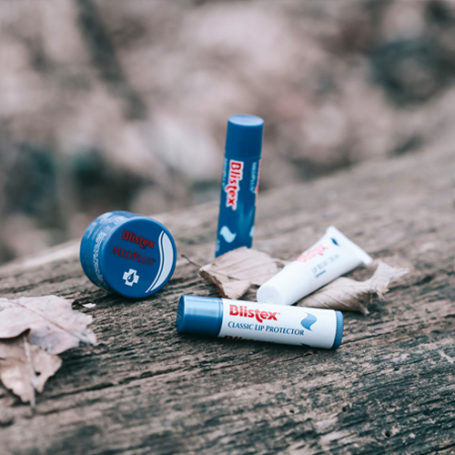 Mehrere Lippenpflegeprodukte von Blistex liegen auf einer Holzfläche mit Herbstlaub daneben.
