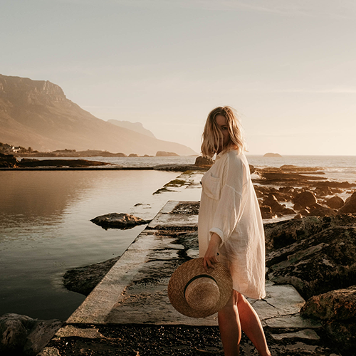 Eine Frau steht in einer sommerlichen Landschaft mit See und Bergen und hält einen Hut in der Hand.