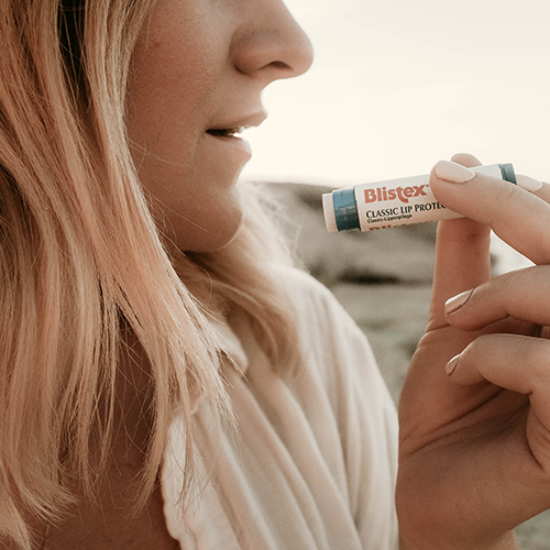 Nahaufnahme eine Frau, die einen Blistex-Lippenpflegestift zu ihren Lippen führt. Sie steht draußen.