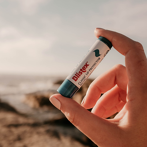 Ein Blistex-Lippenpflegestift wird in der Natur zwischen Daumen und Zeigefinger gehalten.
