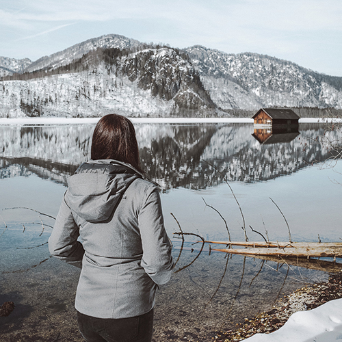 Eine Frau steht an einem Bergsee und schaut sich das verschneite Panorama an.